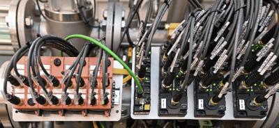Nahaufnahme einer industriellen Anschluss- und Steuerungseinheit mit mehreren schwarzen Kabeln, Steckverbindern und Modulen, die geordnet auf Metallkomponenten montiert sind; gr&uuml;ne Status-LEDs zeigen den Betriebszustand an.