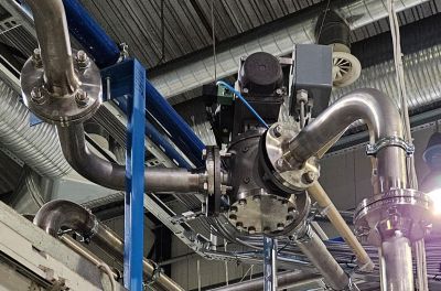 Una válvula de tres vías en un entorno industrial, conectada a un complejo sistema de tuberías soportado por vigas de acero azul. La válvula está equipada con un actuador y componentes eléctricos.