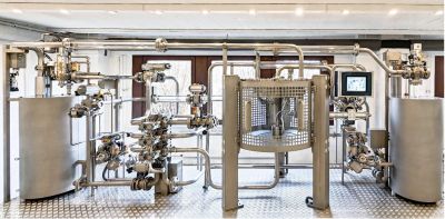 Industrielle Demonstrationsanlage mit mehreren Edelstahltanks, einem komplexen Rohrleitungssystem mit Ventilen und Armaturen sowie einem Touchscreen-Bedienfeld; die Anlage steht in einem hellen Raum mit strukturiertem Bodengitter und zeigt ein vollst&auml;ndiges Molchsystem zur Prozessoptimierung.