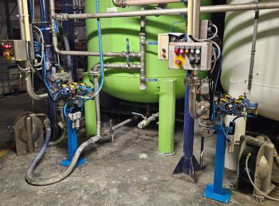Estación de limpieza industrial con recipiente de presión cilíndrico verde, estaciones de operador con paneles de control y un sistema de tuberías complejo con válvulas y actuadores neumáticos en una planta de producción.