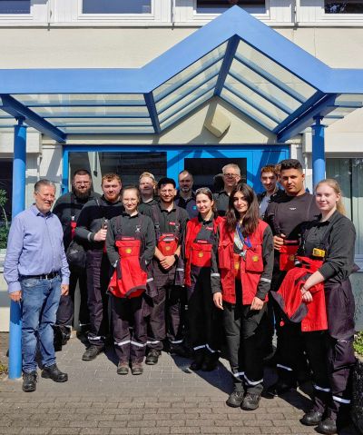 Gruppenfoto vor dem I.S.T. Gebäude-Eingang: Eine größere Gruppe von Personen in schwarzer Arbeitskleidung mit roten Schutzwesten steht unter einem blauen Glasvordach und blickt in die Kamera. Einige halten rote Schutzkleidung in der Hand; sonniges Tageslicht.